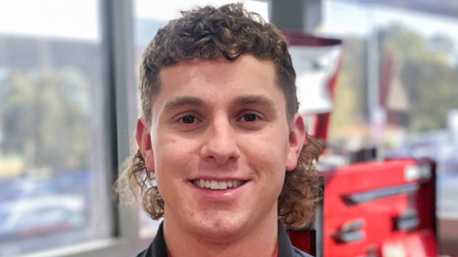 A young man with blonde hair, smiling, wearing a red Toyota mechanic shirt, inside of a Toyota dealership.