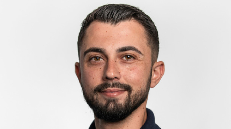 A content looking man with black hair and a modest beard, wearing a black polo shirt in front of a white backdrop.