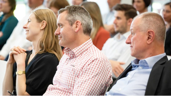 A large crowd of women and men in a naturally-lit indoor environment enjoying a presentation attentively.