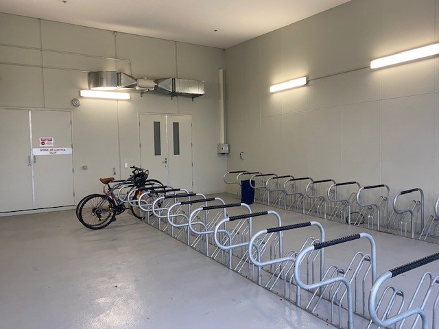 An indoor bicycle parking area with multiple metal bike racks, a single bike parked, with white walls and fluorescent lighting