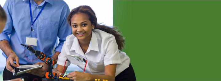 A young man and woman smiling, testing a small mechanical project. The man is holding a notepad, whilst the woman is connecting wires under a magnifying glass.
