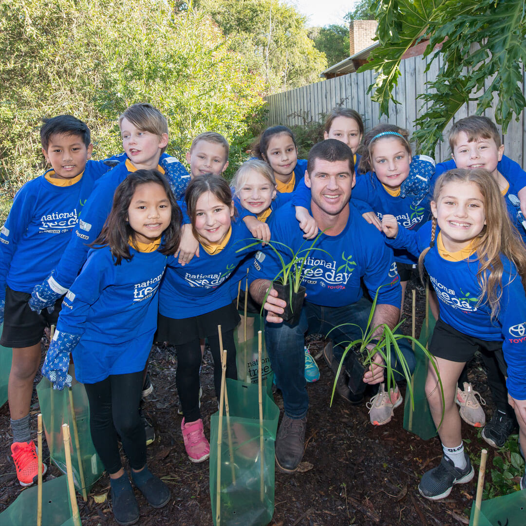 A group of children and former AFL player Jonathan Brown holding potted saplings in his hands, crouching down for a group photo on a sunny afternoon, surrounded by freshly planted trees, all wearing blue National Tree Day jumpers.