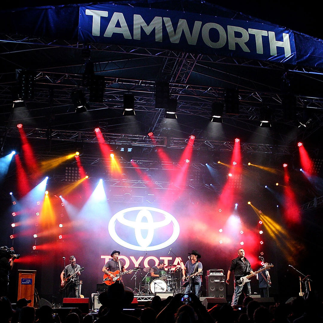 A live performance by a group at the Toyota Tamworth Country Music Festival, red and orange spotlights shining onto the stage and the crowd, with a giant glowing Toyota logo lit up behind the performers.