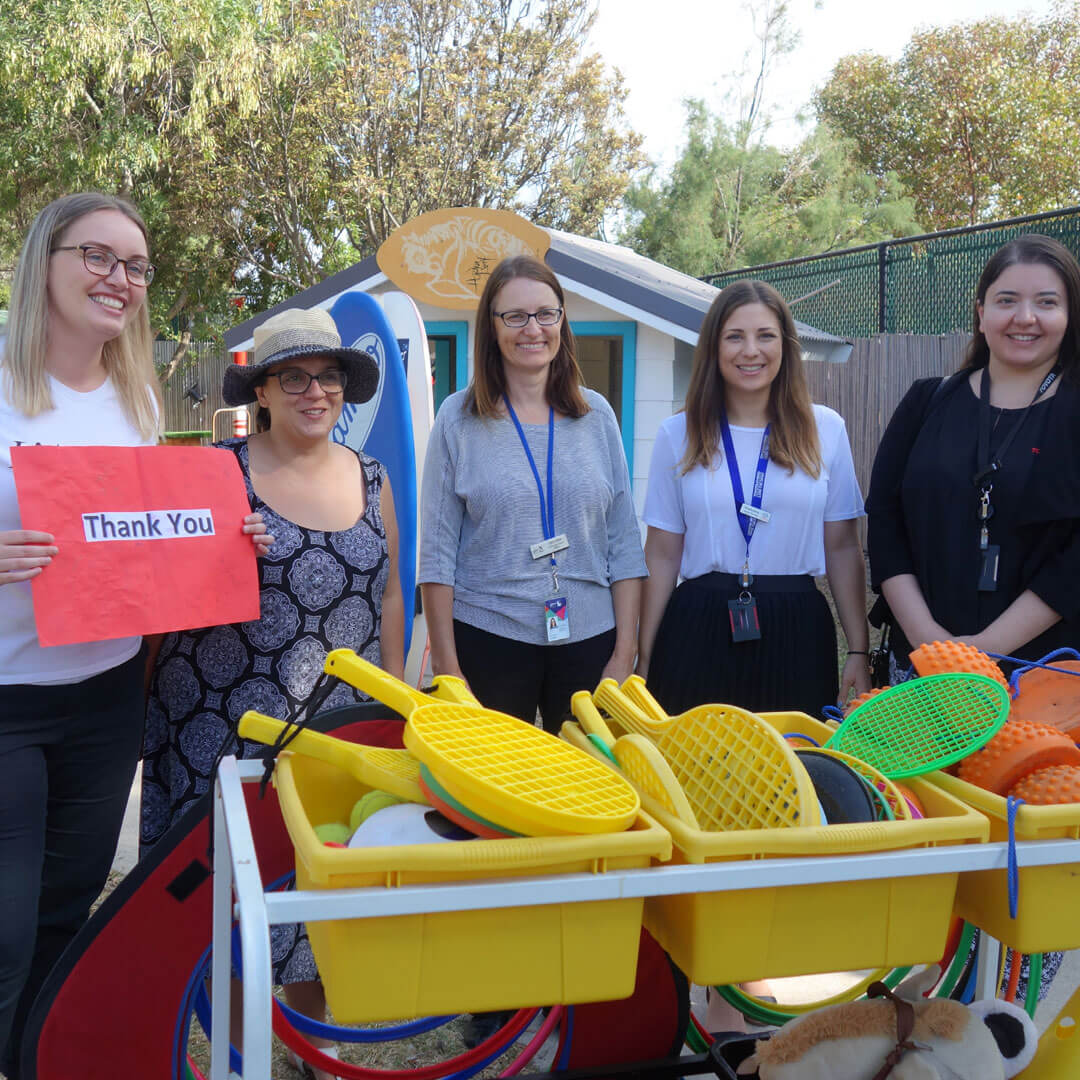 Five women with a ‘Thank You’ sign held up, surrounding yellow tubs of donated children’s sports gear such as tennis rackets, tennis balls, sports cones and hula hoops.