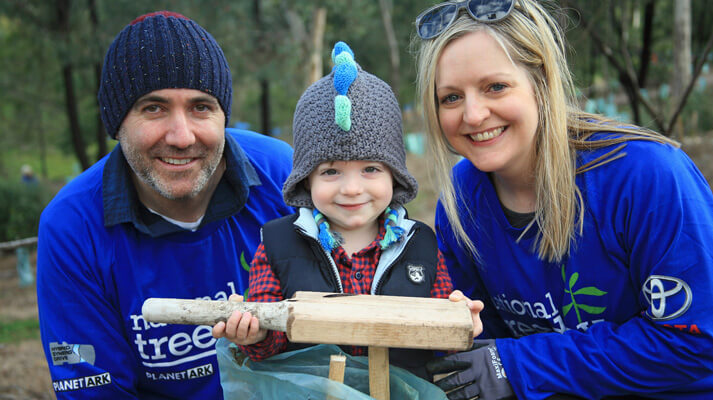 Two adults and a toddler, smiling contently wearing blue National Tree Day shirts, the toddler grinning cheekily, holding a wooden hammer used to knock in the posts that support a freshly planted tree.