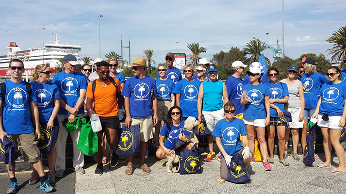 A group of people wearing blue Port Melbourne Beach Patrol shirts, posed together at Port Melbourne during the day, with the Spirit of Tasmania docked in the distance behind them.