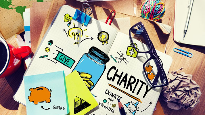 A bird’s eye view of a notebook amongst a scattered desk surrounded by papers, a pair of reading glasses, paperclips, a ball of rubber bands, pens, and a coffee mug. On the notebook is creatively written: “Give”, “Charity”, “Donate”, “Help” and “Volunteer”.
