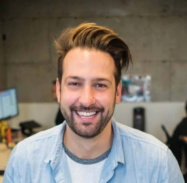 A man with brown hair in a quiff haircut, smiling happily under warm lighting, wearing a washed out denim shirt, in a modern industrial office environment with an exposed concrete wall in the background.