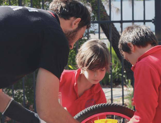 A man and two young children outside at a primary school, working together to repair the wheel on a bicycle.