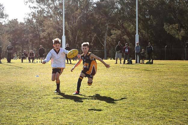 Two young primary school aged boys wearing opposing football uniforms, chasing a yellow football that’s bouncing midair during a sunny afternoon, with adults watching from afar.