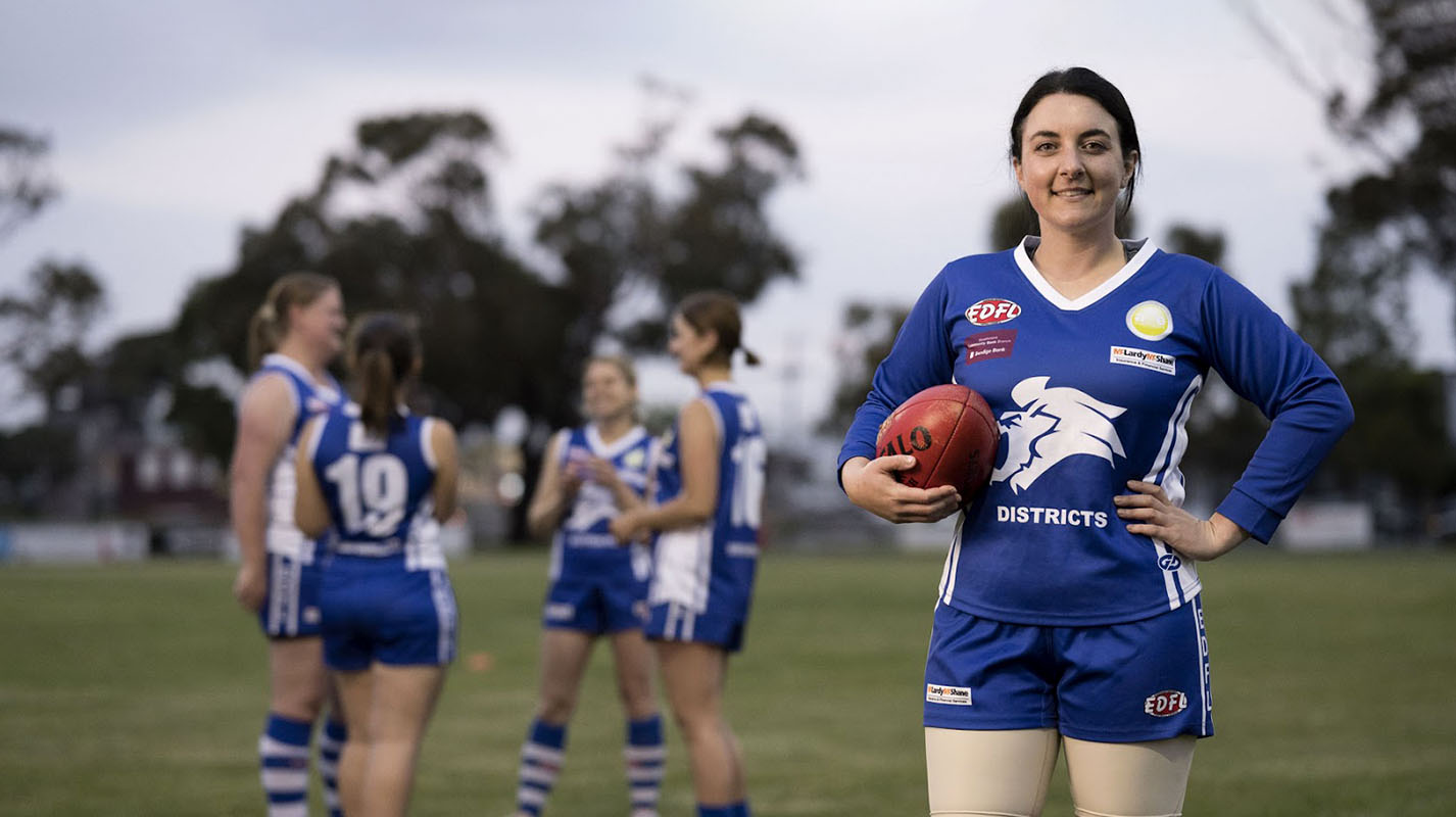 An AFLW player standing on a footy oval, smiling proudly, holding a football at her side, wearing a blue and white footy uniform. Her teammates in the background.