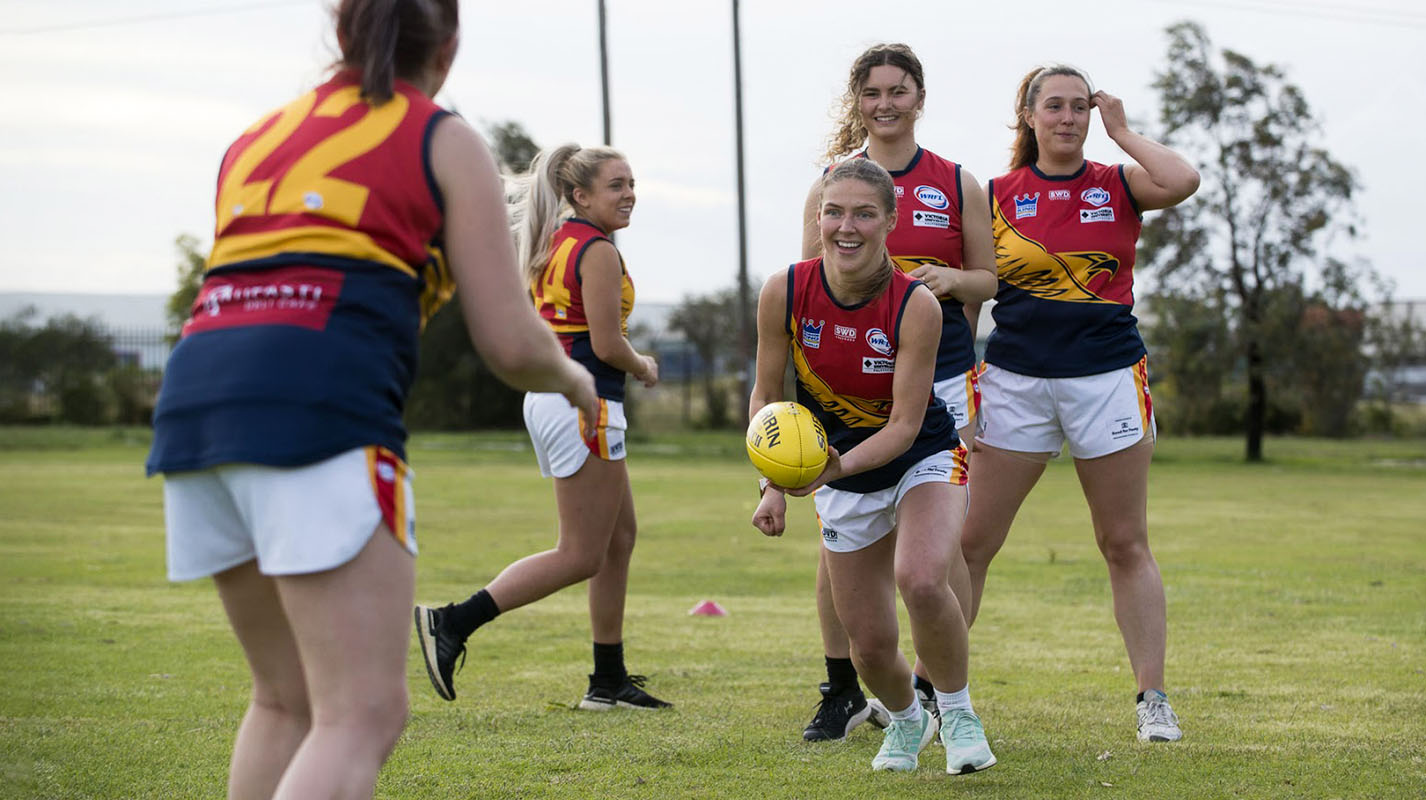 AFLW players enjoying footy practice on an oval, one about to punt a yellow football to another fellow team member.