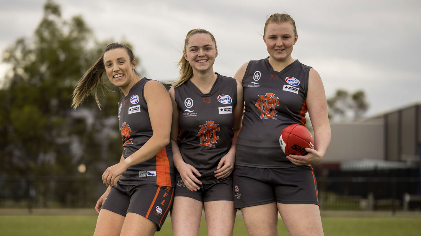 Three youthful AFLW players wearing black and red uniforms together next to each other on a footy oval, each sporting a grin.