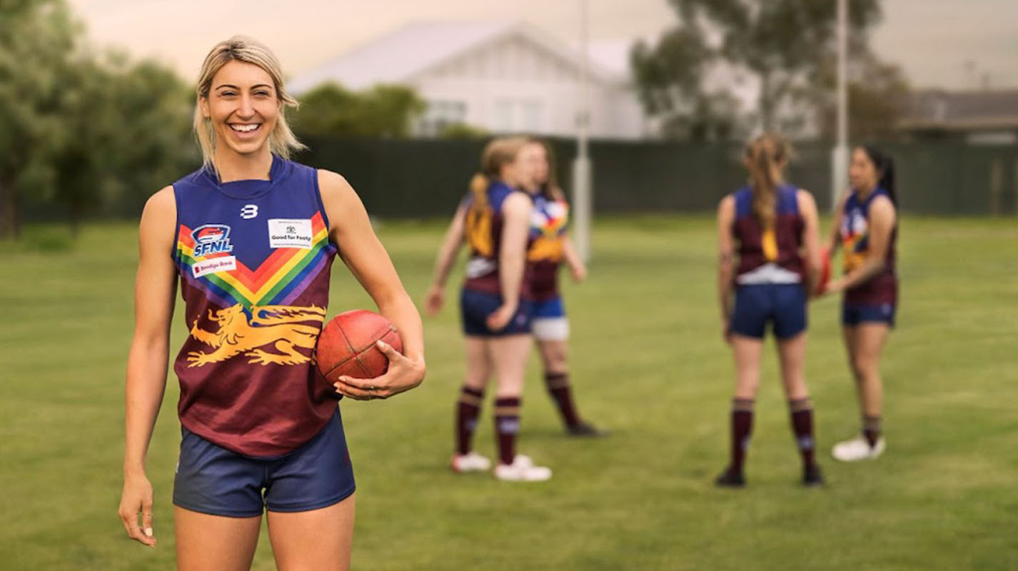 An AFLW player standing on a footy oval, happily smiling holding a football at her side, wearing a Brisbane Lions jersey featuring a rainbow design. Her teammates in the background.