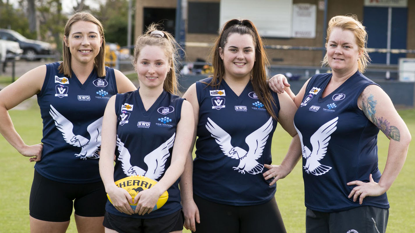 Four AFLW players, standing next to each other on a footy oval in solidarity, wearing a dark navy and white footy uniform.