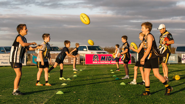 A kids footy team, practicing with their footy coach on a sunny afternoon, standing in adjacent lines and punting balls to one another.