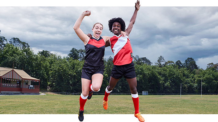 Two football players, one arm around each other, happily jumping up in the air on a footy oval, each raising one fist towards the sky.