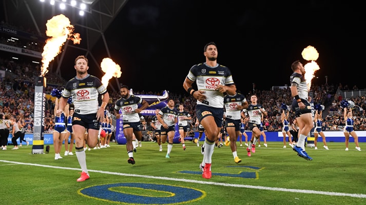 The rugby league team, North Queensland Cowboys running across a field during a night match under beaming lights, flames shooting from pillars overhead.