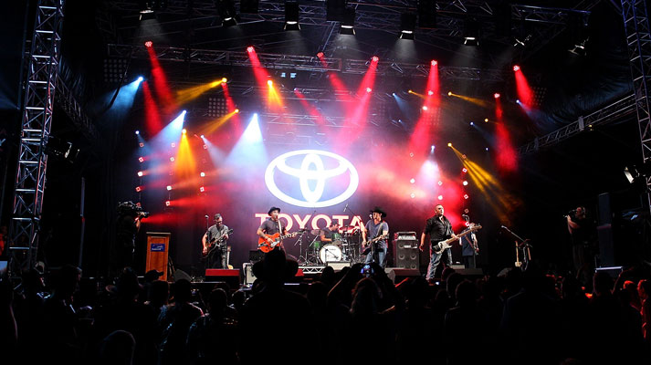 A live performance by a group at the Toyota Tamworth Country Music Festival, red and orange spotlights shining onto the stage and the crowd, with a giant glowing Toyota logo lit up behind the performers.