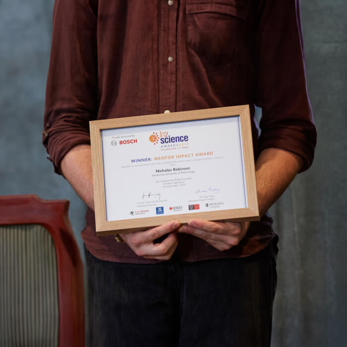 A person in a burgundy shirt holds up an ‘In2science’ Mentor Impact Award.