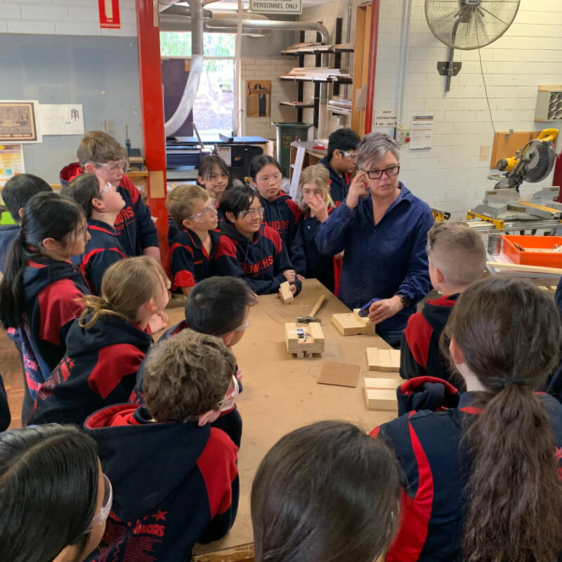 A class of children in navy blue and red school uniforms, sitting around a table guided by a teacher in a neatly organised wood workshop, as a part of the Schools Plus program.