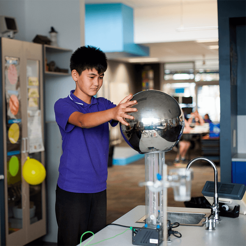 A young schoolboy in a classroom wears a purple polo shirt uniform. He is touching a Van de Graaff generator with both hands, his thick black hair standing on end from the static electricity coursing through his body.