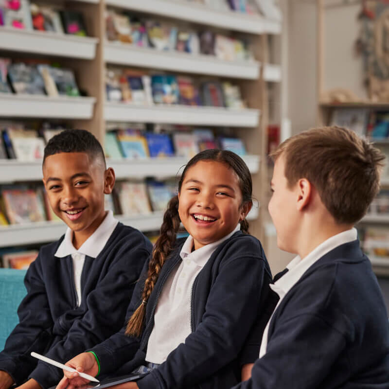 Three uniformed school children in a school library environment sit together and laugh.