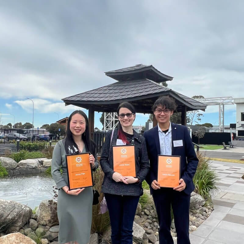 Three people stand together proudly holding up Western Chances certificates, in front of a pavilion reminiscent of East-Asian architecture, on a cloudy day.