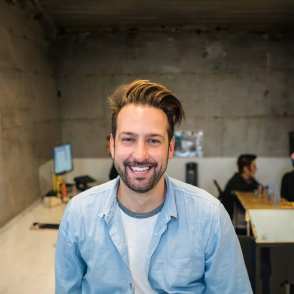 A man with brown hair in a quiff haircut, smiling happily under warm lighting, wearing a washed-out denim shirt, in a modern industrial office environment with an exposed concrete wall in the background.