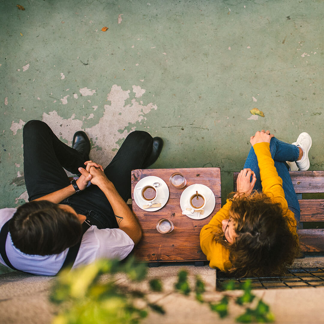 A top-down view of a woman and a man sitting together in an outdoor environment conversing, a small wooden table between them, placed on the table is a coffee and a glass of water for each of them.