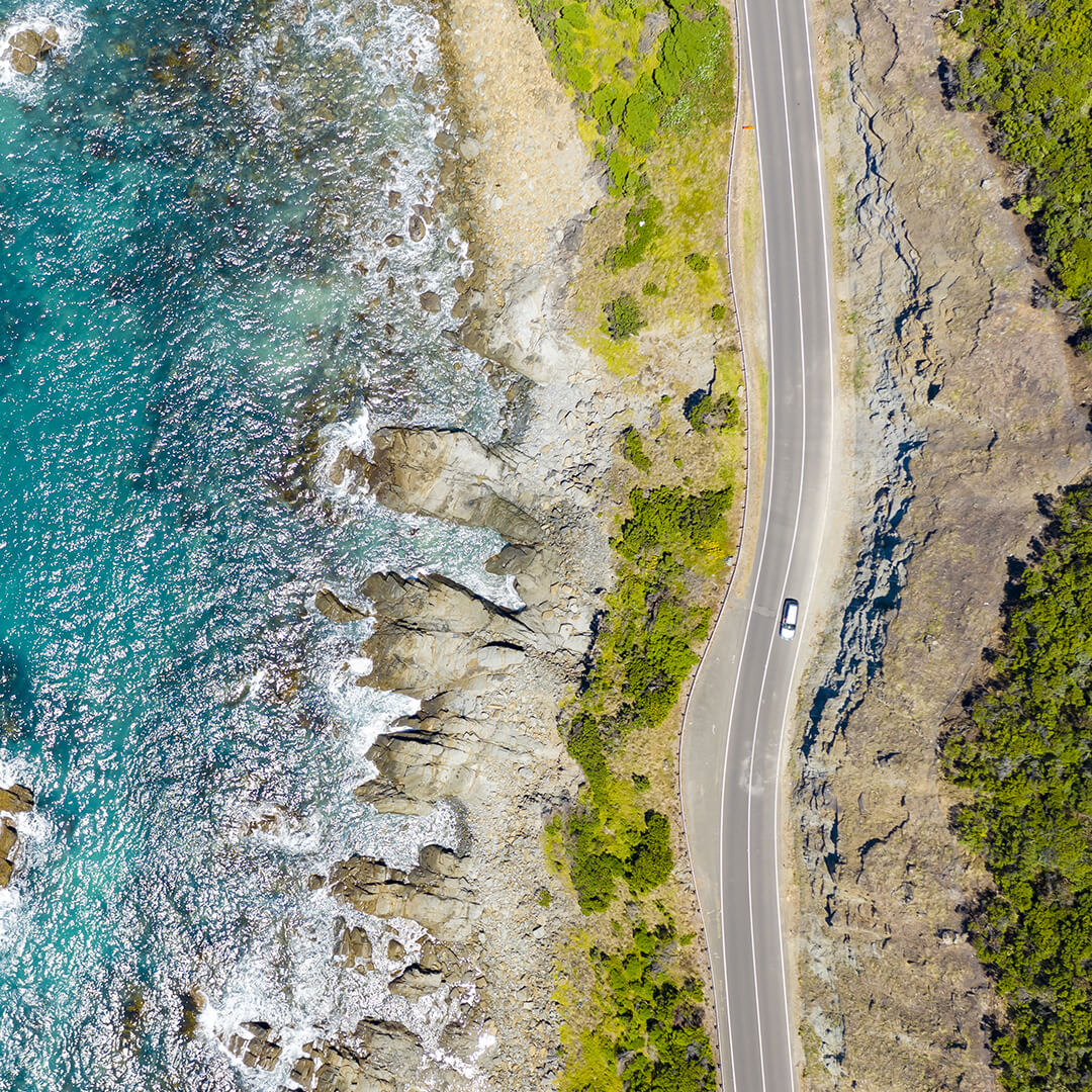 A birds-eye view of a rocky coast on a clear day; to the left, pristine blue ocean with seafoam crashing upon the rocks, to the right a curved highway bordered with vivid green shrubbery, and on that road a silver car travelling south.