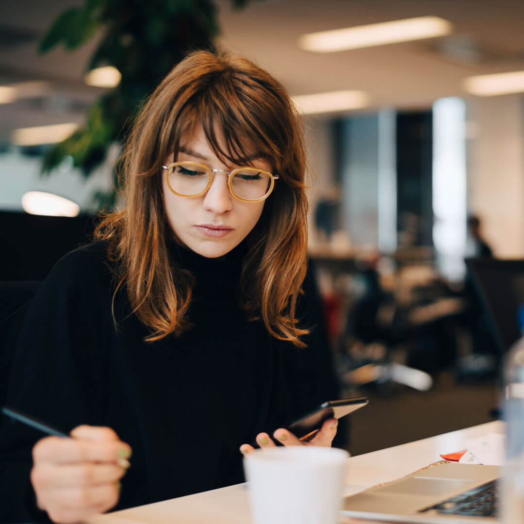 A woman with rich wavy natural, brown and caramel hair, with distinct winged eyeliner, wearing large yellow reading glasses and a black turtleneck, reading, her attention turned downwards, in a warmly lit office environment blurred out in the background.