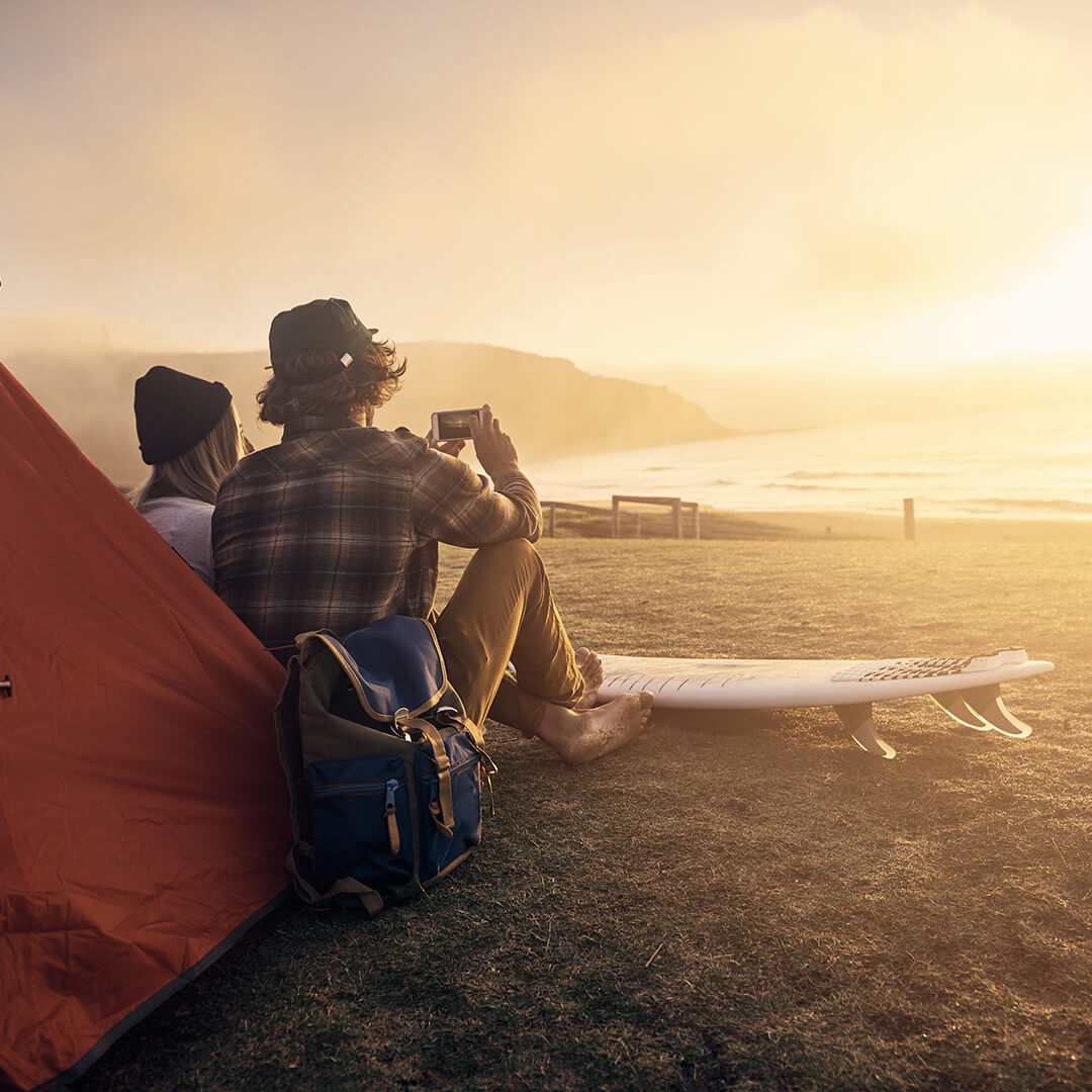 A view of a barefooted man and a woman sitting closely together in front of a tent on a beach, a backpack and a surfboard nearby, jointly taking a photo of the brilliant sun setting over a calm ocean
