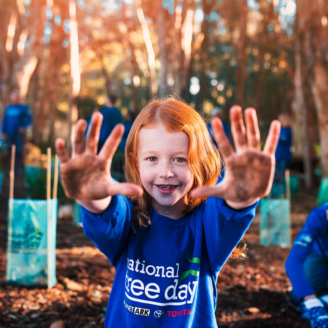 A young child with vivid orange shoulder-length hair, lots of freckles, and hazel eyes, smiling cheekily, wearing a bright blue National Tree Day jumper with their arms outstretched and their dirt-covered hands held up towards the viewer, surrounded by freshly planted trees in the Australian bush on a warm sunny afternoon.