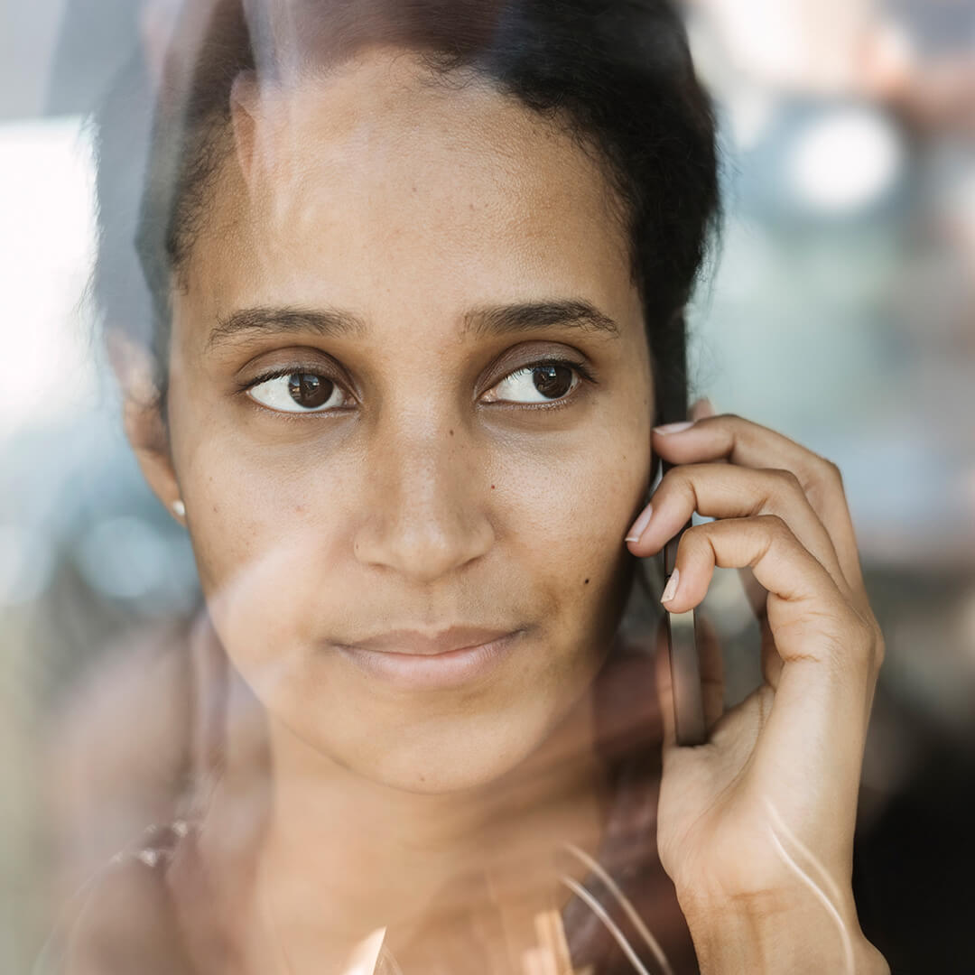 The close-up of a woman with dark brown hair and deep brown eyes, on a mobile phone behind a pane of glass, looking out into the distance, a pensive expression on her face.
