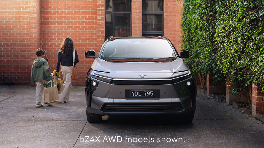 A bZ4X in Frosted White drives along a sunlit highway bordered by trees and open fields. The motion blur emphasizes its smooth movement, with text at the bottom reading, Overseas model shown.