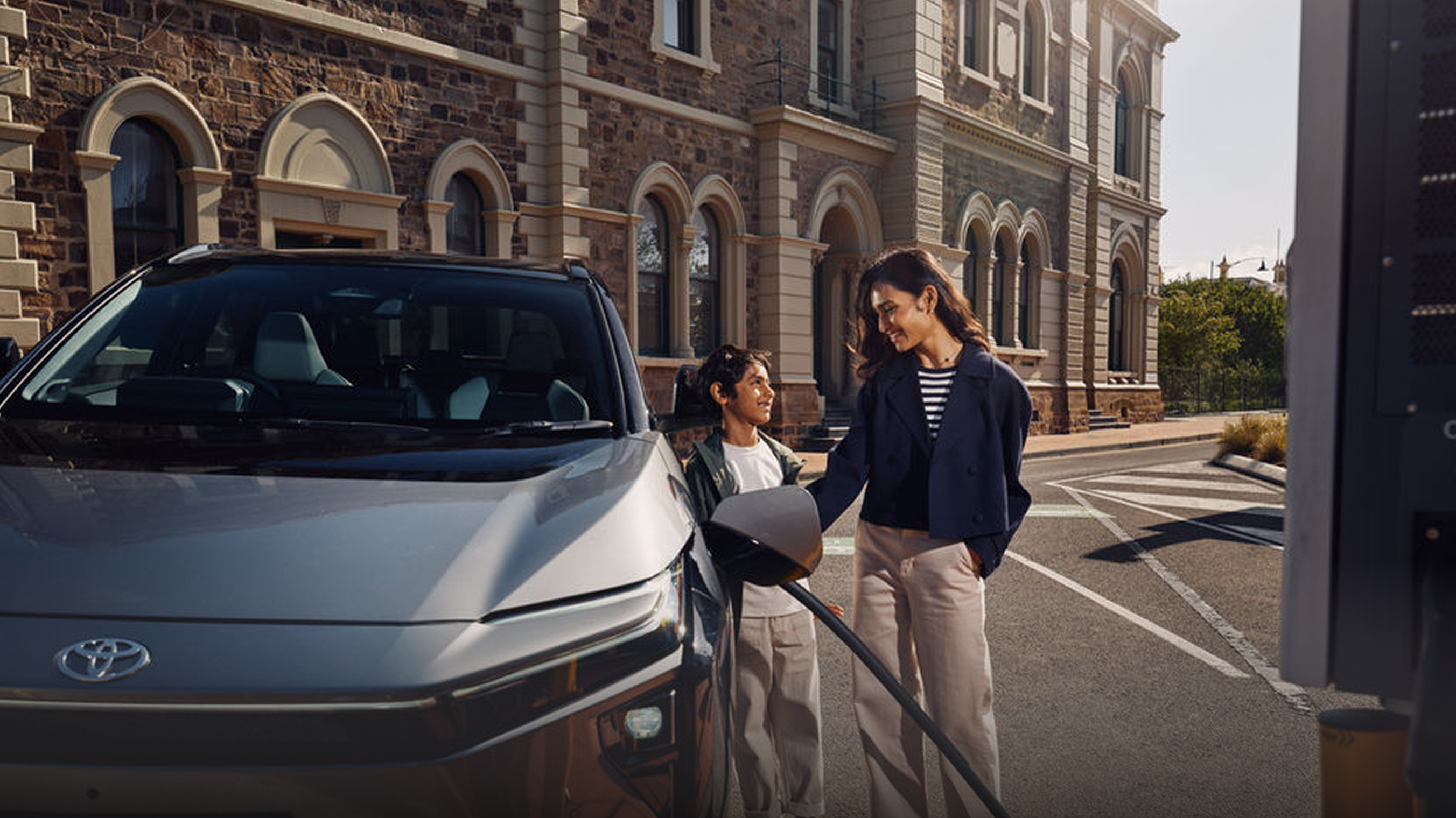 A woman and a child stand beside a gray electric bZ4X that’s plugged in and charging on a city street. They smile at each other while the car charges, with stone building in the background.