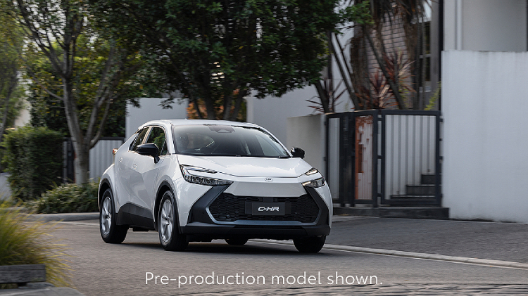 A C-HR in Frosted White paint drives through a quiet suburban street lined with trees and modern homes. The compact SUV features sharp, angular styling and LED headlights. Text at the bottom reads, Overseas model shown.