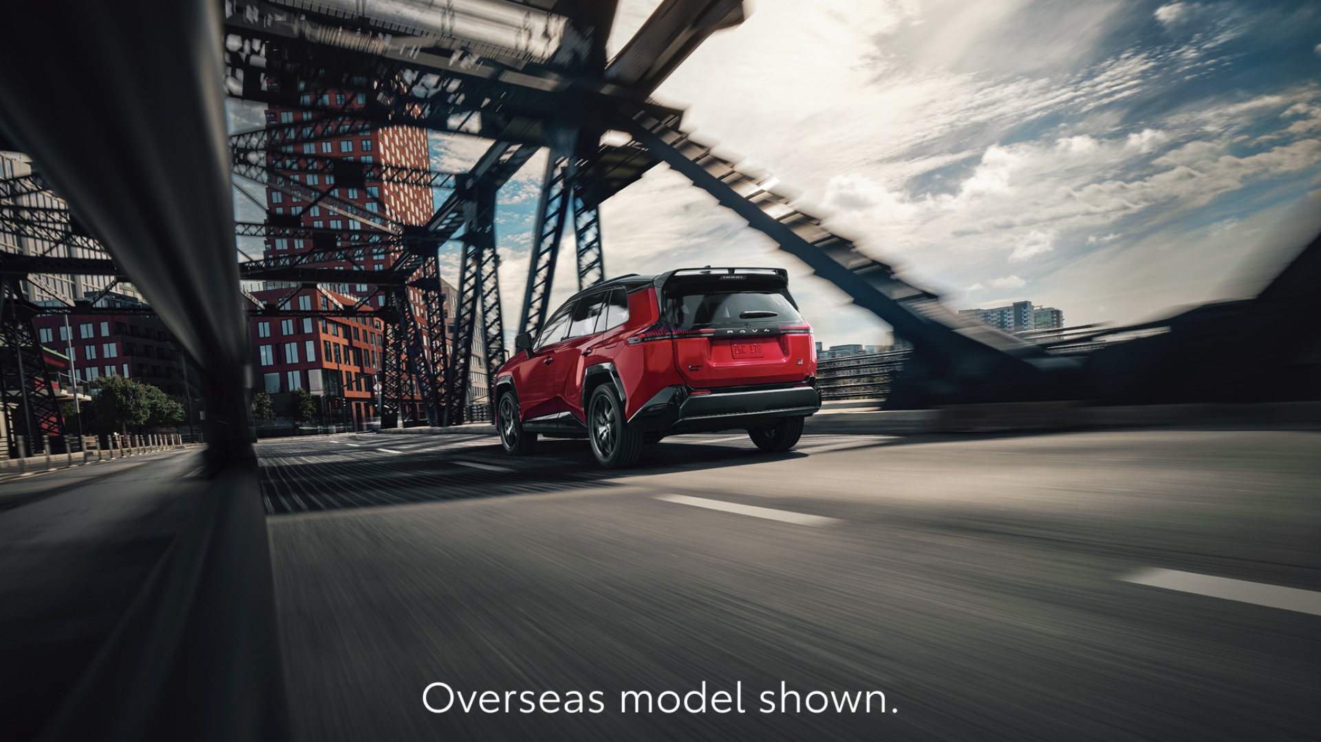 A RAV4 in Feverish Red paint drives across a steel bridge in an urban cityscape under a partly cloudy sky. The image captures the rear and side view of the SUV in motion, with the text, Overseas model shown displayed at the bottom.