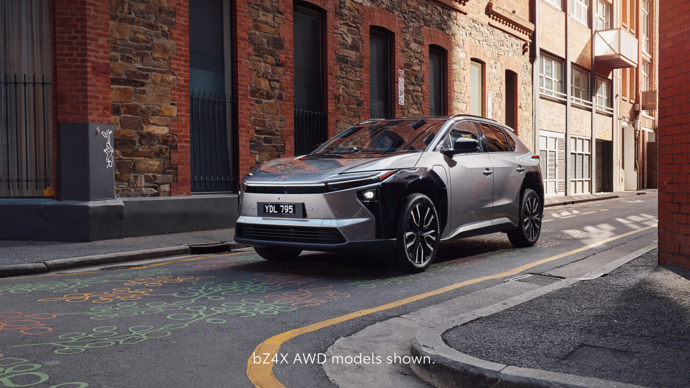 A bZ4X in Frosted White is parked beside a modern concrete and metal building, viewed from the side to show its sleek, angular design and black wheel arches. Text at the bottom reads “Pre-production models shown.”