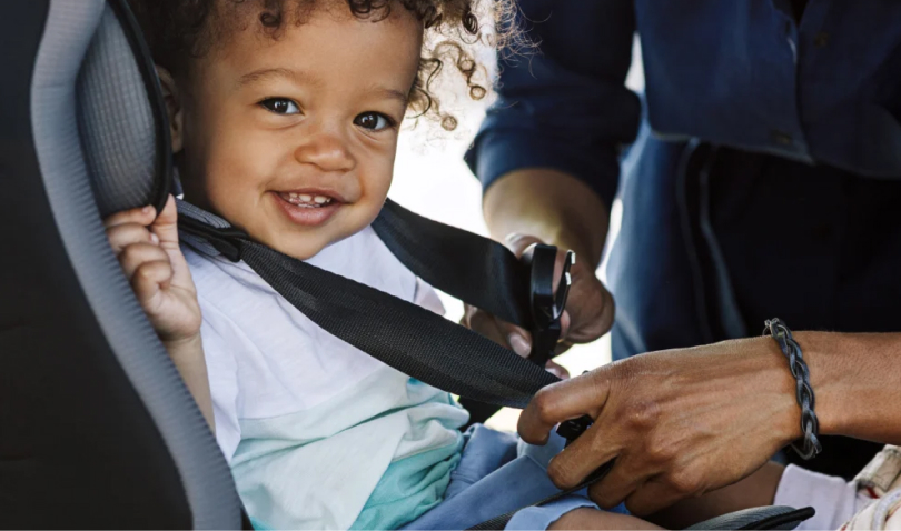 A smiling toddler sitting in a car seat while an adult fastens their seatbelt.