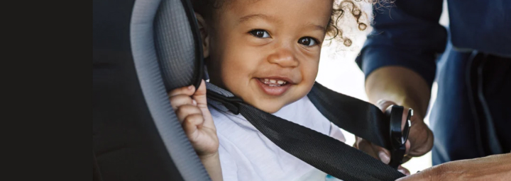 A smiling toddler sitting in a car seat while an adult fastens their seatbelt.