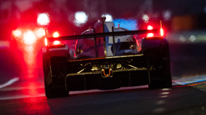 The back of a Formula 1 racing vehicle in the rain at night, with red lighting scattered off in the distance.