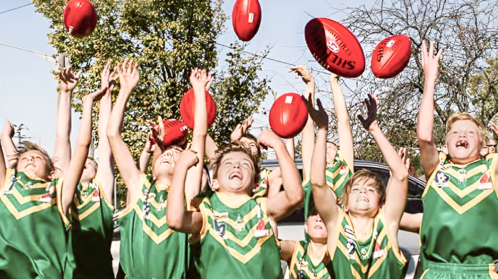A group of young boys in green and yellow football jerseys, smiling and throwing red Sherrin footballs up into the air.
