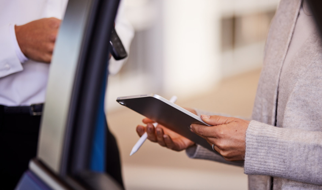 A woman stands next to a Toyota Dealer, holding a digital tablet and accompanying pen, the door of a blue Toyota ajar.