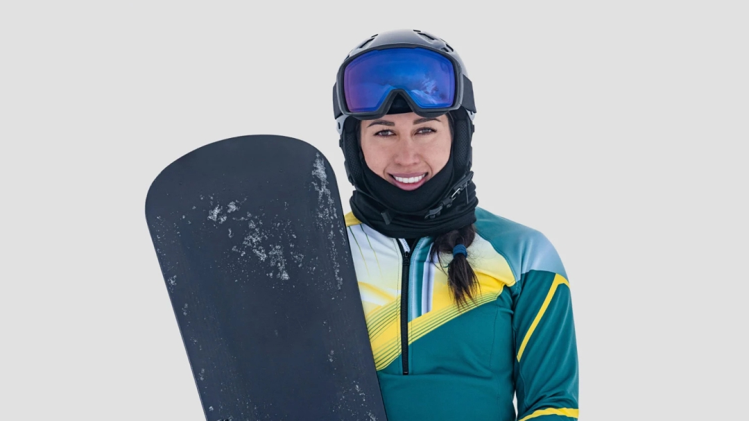 Australian Olympian snowboarder Belle Brockhoff stands smiling, fully-dressed in snowboarding gear, holding a snowboard in her right arm.
