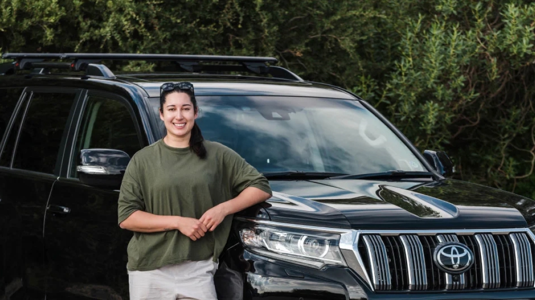 Australian Olympian snowboarder Belle Brockhoff in a lush bushland area, smiling as her left arm rests on the bonnet of her black LandCruiser Prado.