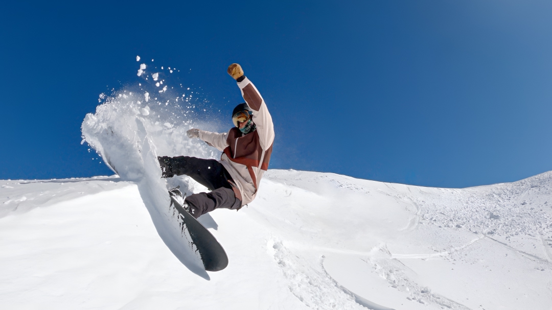 Australian Paralympian snowboarder Ben Tudhope, kicking up snow mid-air, while snowboarding down a mountain.