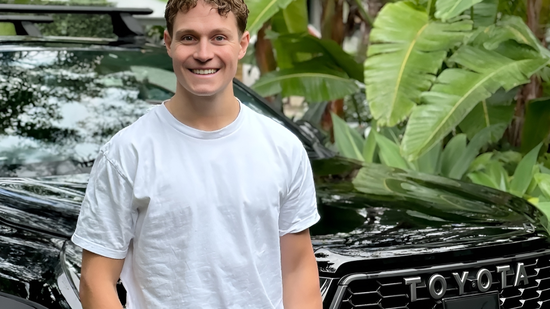 Australian Paralympian snowboarder Ben Tudhope smiling, standing in front of his HiLux GR Sport amongst luscious tropical plantlife.