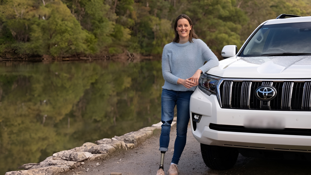 Australian Paralympian swimmer Ellie Cole standing relaxed against her white LandCruiser Prado, parked next to a calm river in the Australian outback.